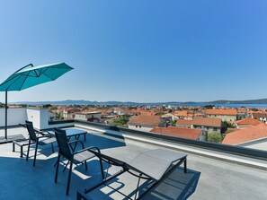 A rooftop terrace with a table and chairs, a blue umbrella, and a view of a coastal town. - Apartment  by Interhome (Zadar/Bibinje)