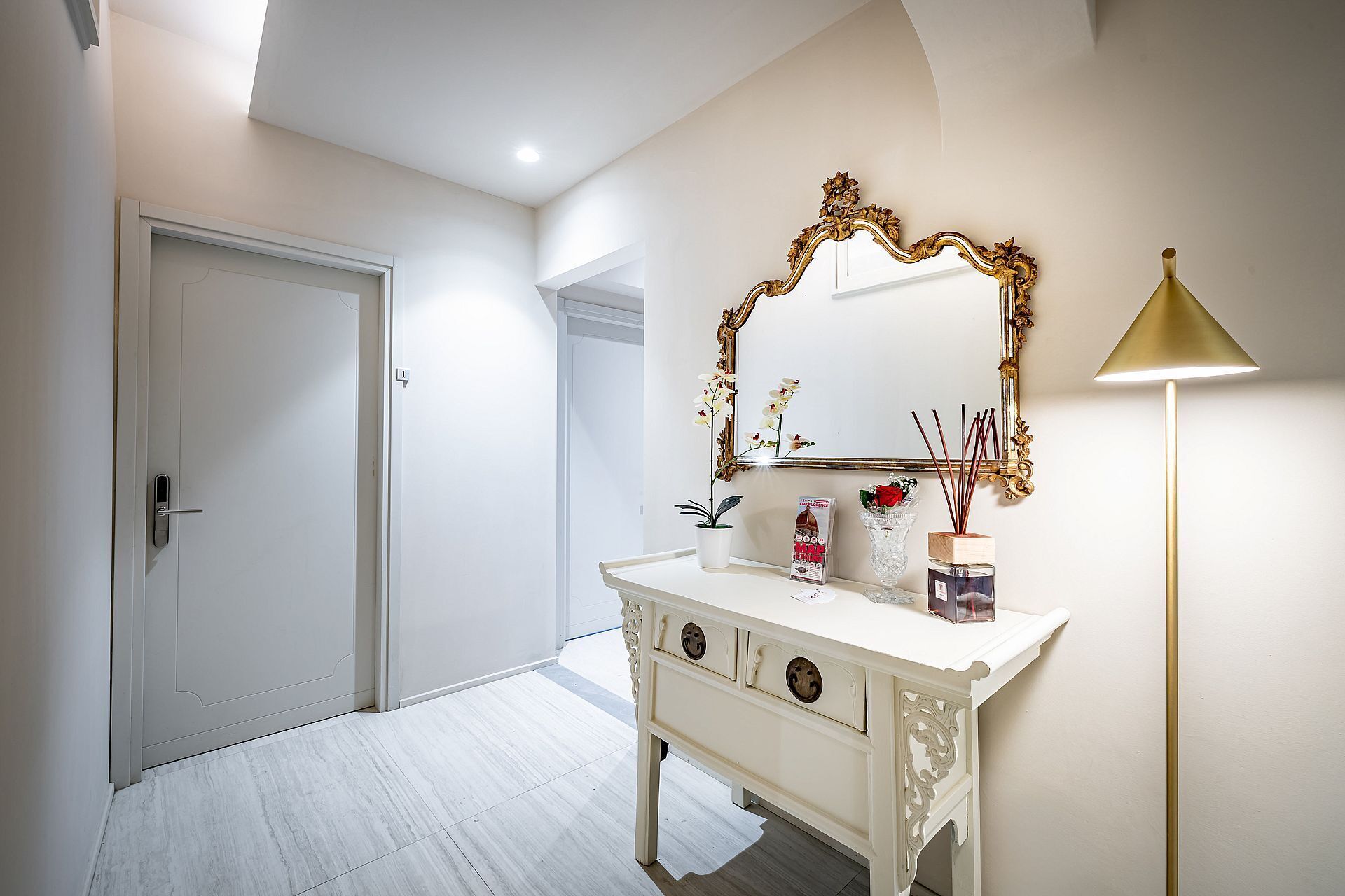A bathroom with a white vanity, a mirror with an ornate frame, a gold lamp, and a potted plant.