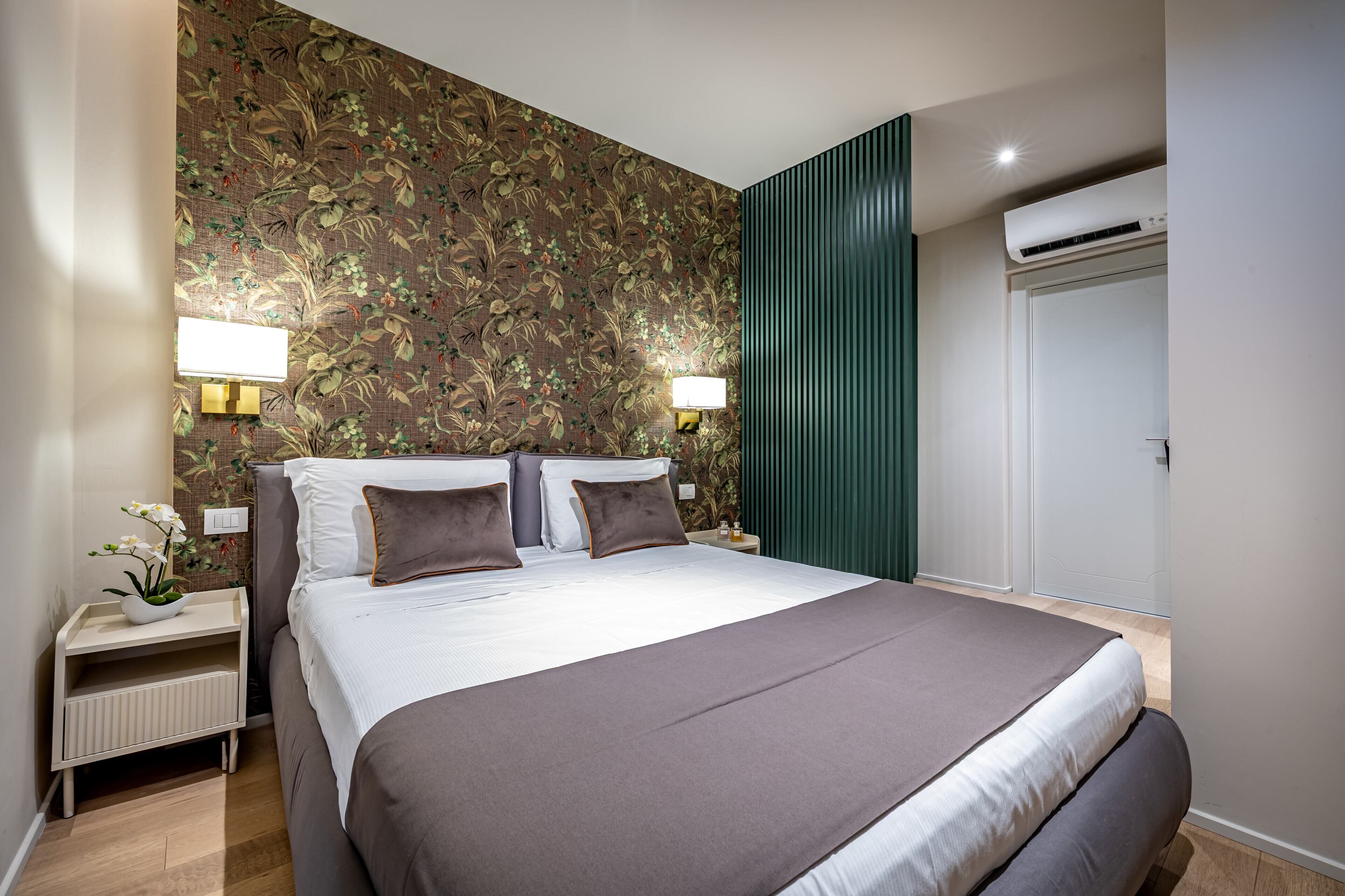 Una cama bien hecha con una colcha marrón y almohadas, una mesita de noche con una lámpara, un papel tapiz floral y un equipo de aire acondicionado.