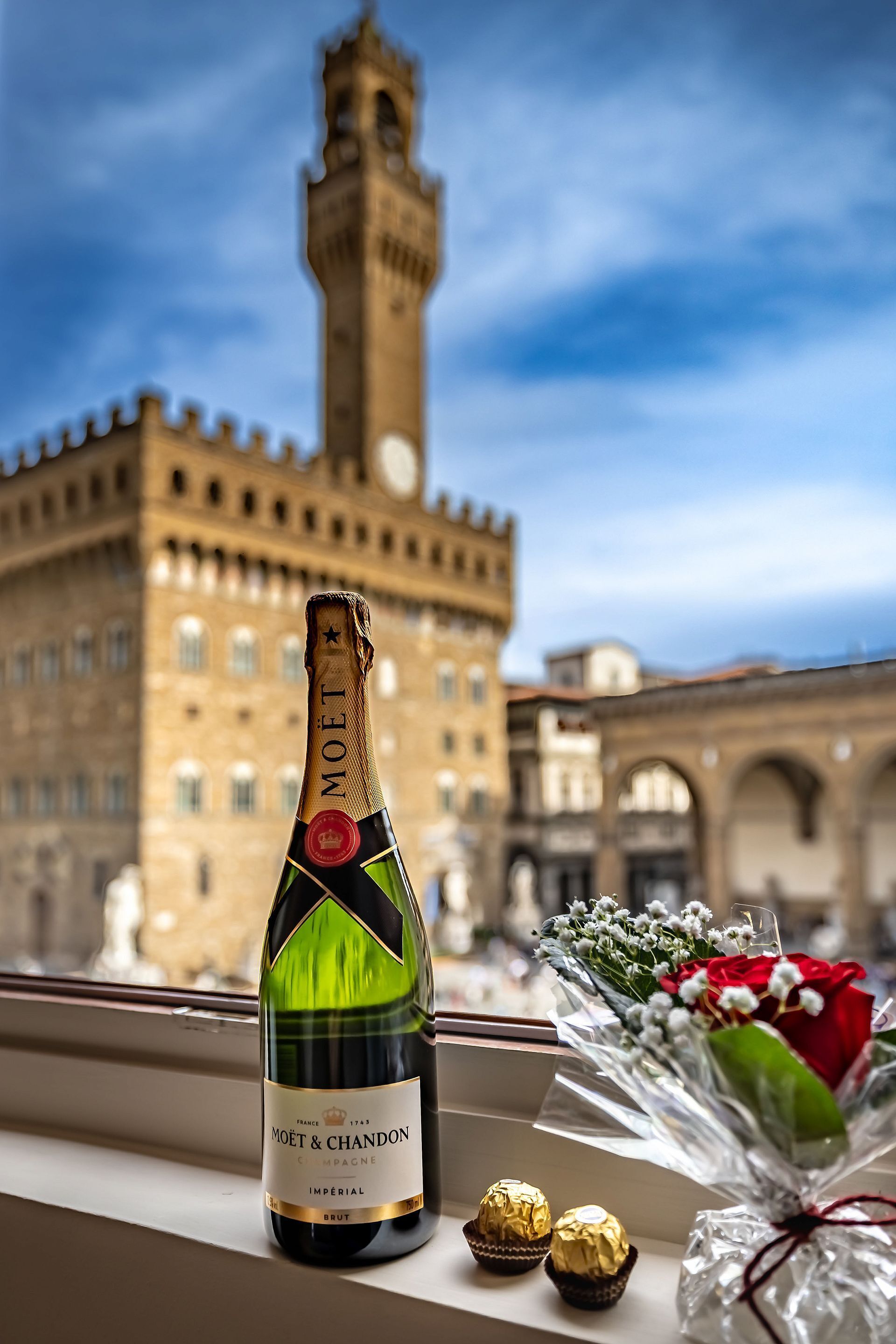 A bottle of Moët & Chandon champagne, a bouquet of flowers, and chocolates on a windowsill with a historic building in the background.