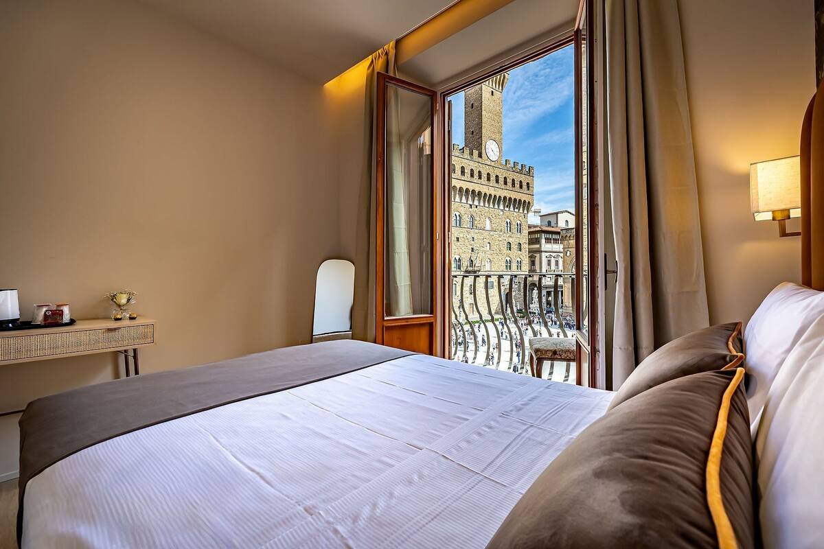 Une chambre d’hôtel comprenant un lit, une table de chevet, un miroir et une fenêtre donnant sur un bâtiment historique.