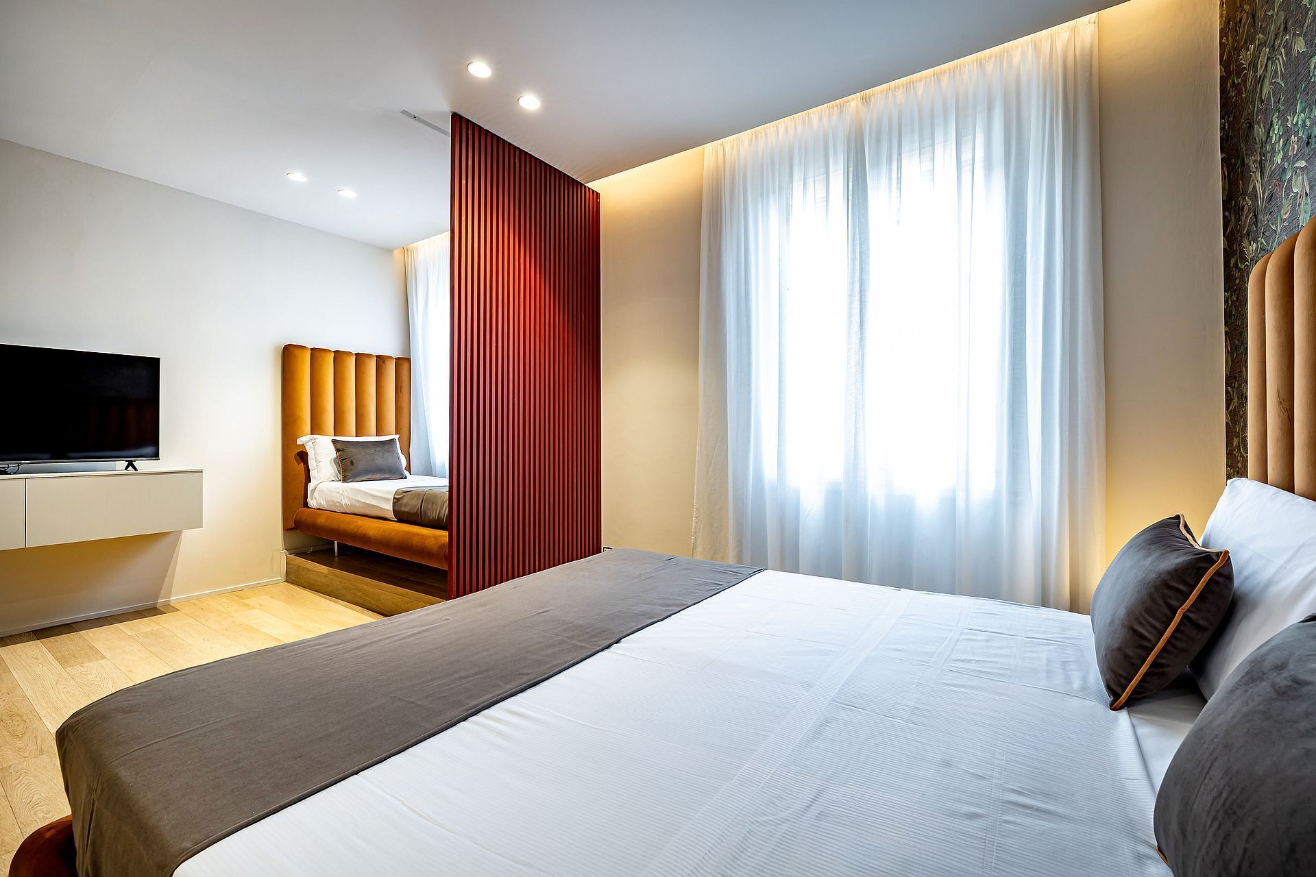 Quarto de hotel moderno com uma cama grande, televisão de tela plana, cabeceira de madeira e uma janela ampla com cortinas leves.