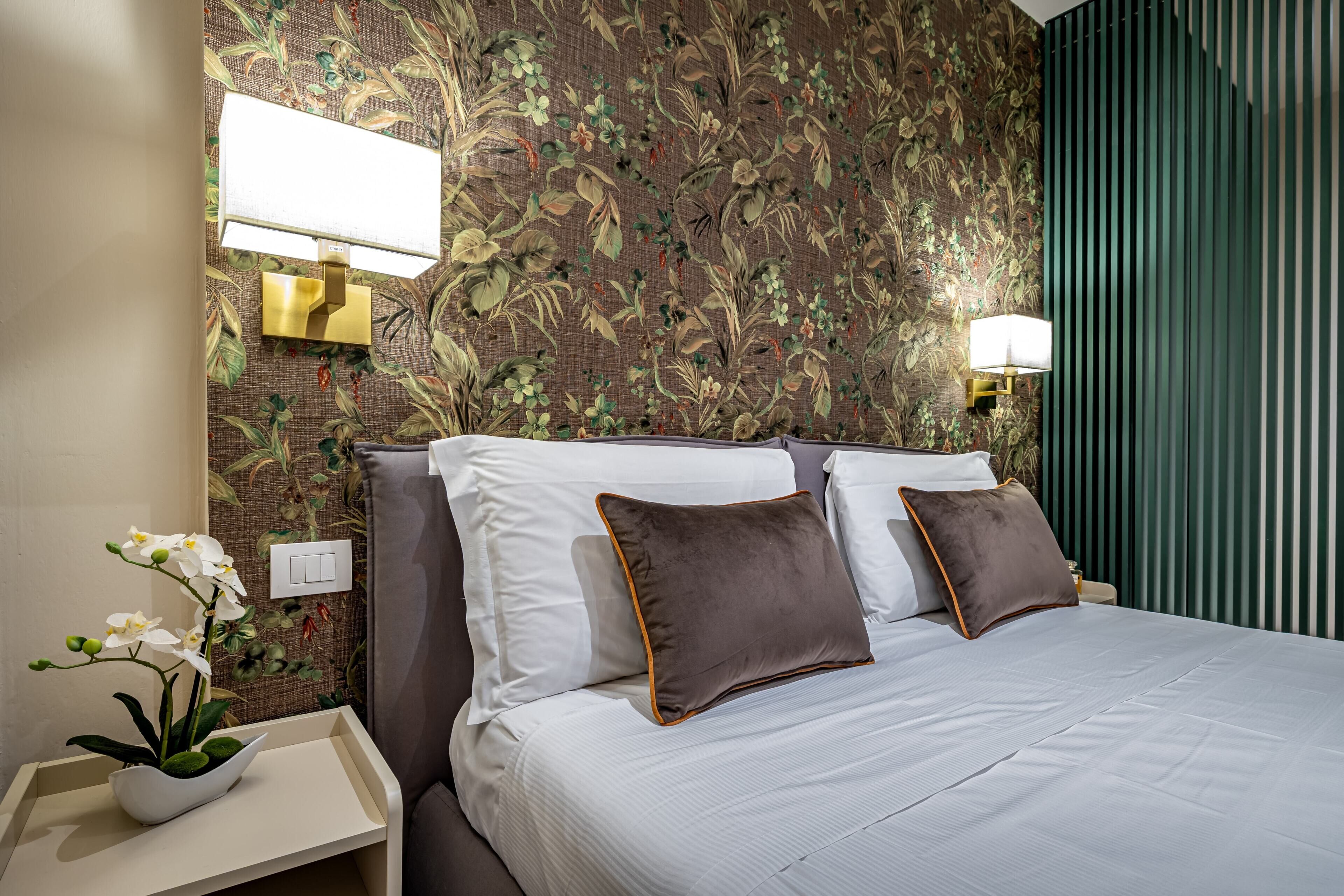 A hotel room with a bed, two wall-mounted lamps, a floral wallpaper, and a nightstand with a plant.