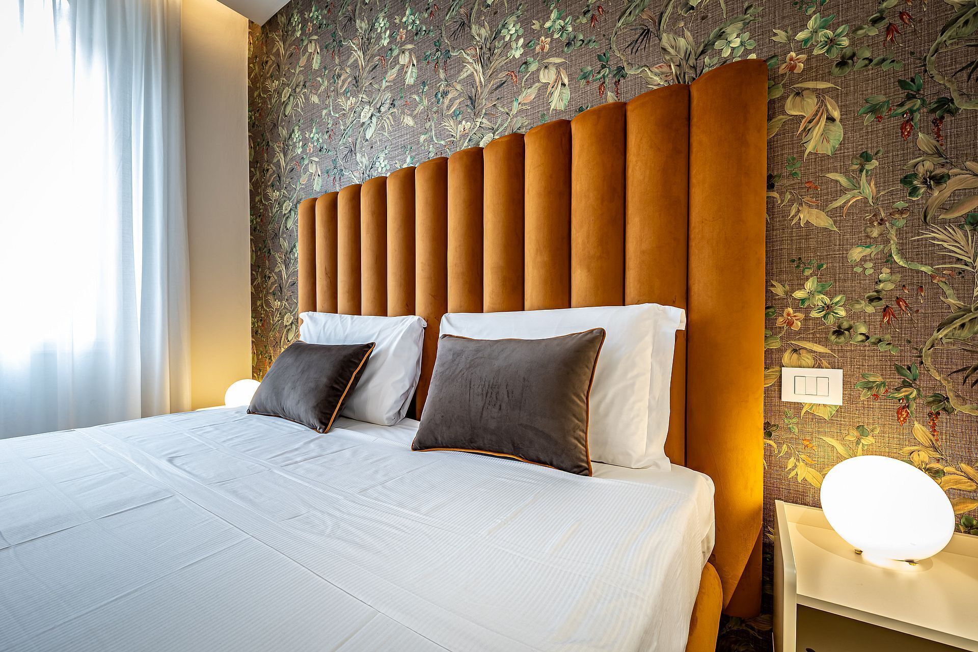 A hotel room with a large bed, a headboard with vertical panels, floral wallpaper, and a bedside table with a lamp.