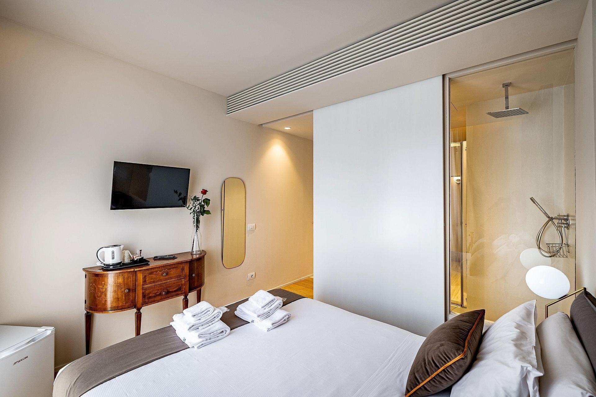 A hotel room with a bed, a TV, a mirror, a wooden side table with a kettle, and a shower area.