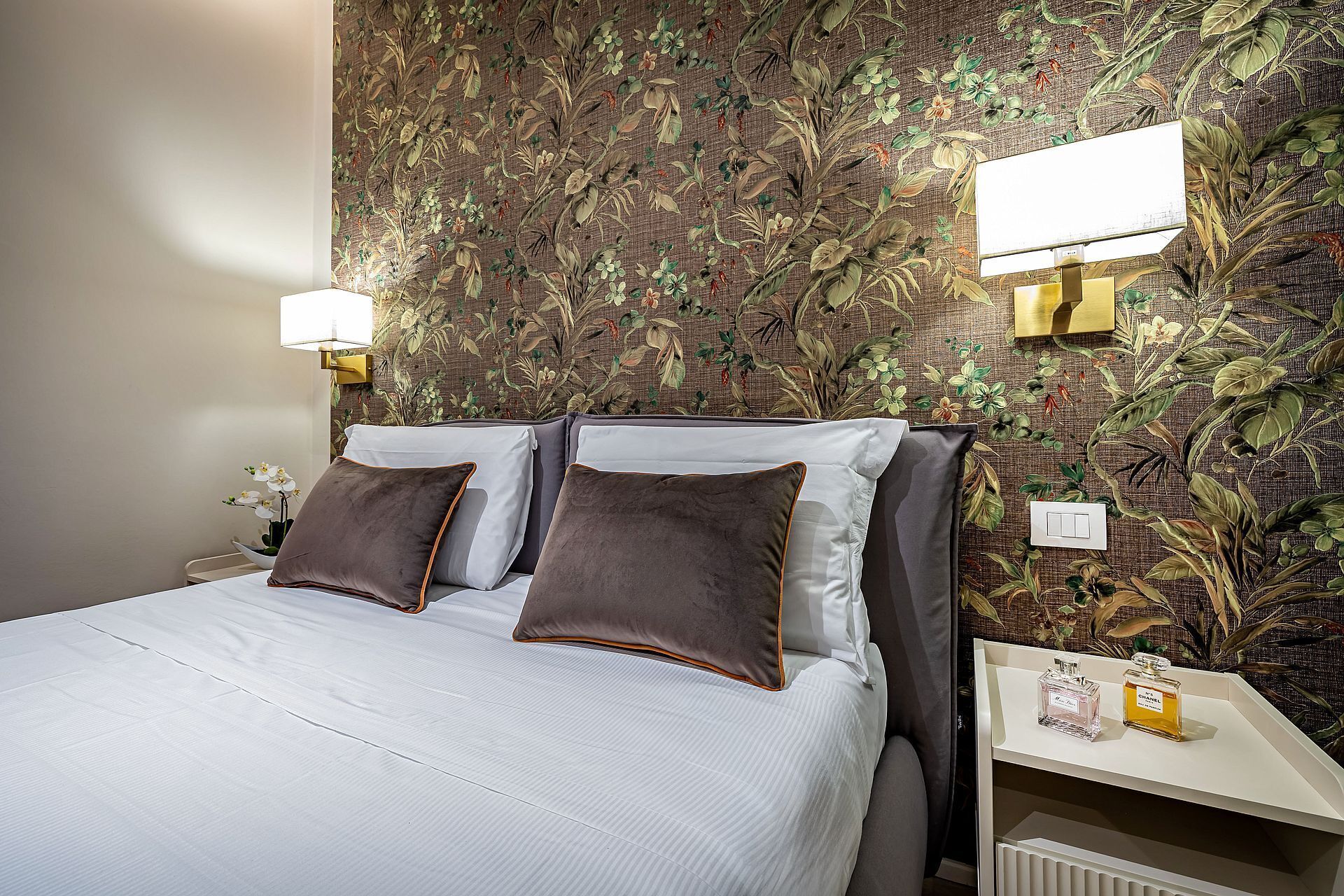 Una cama bien hecha con almohadas, una mesita de noche con una lámpara y una pared con estampado floral.