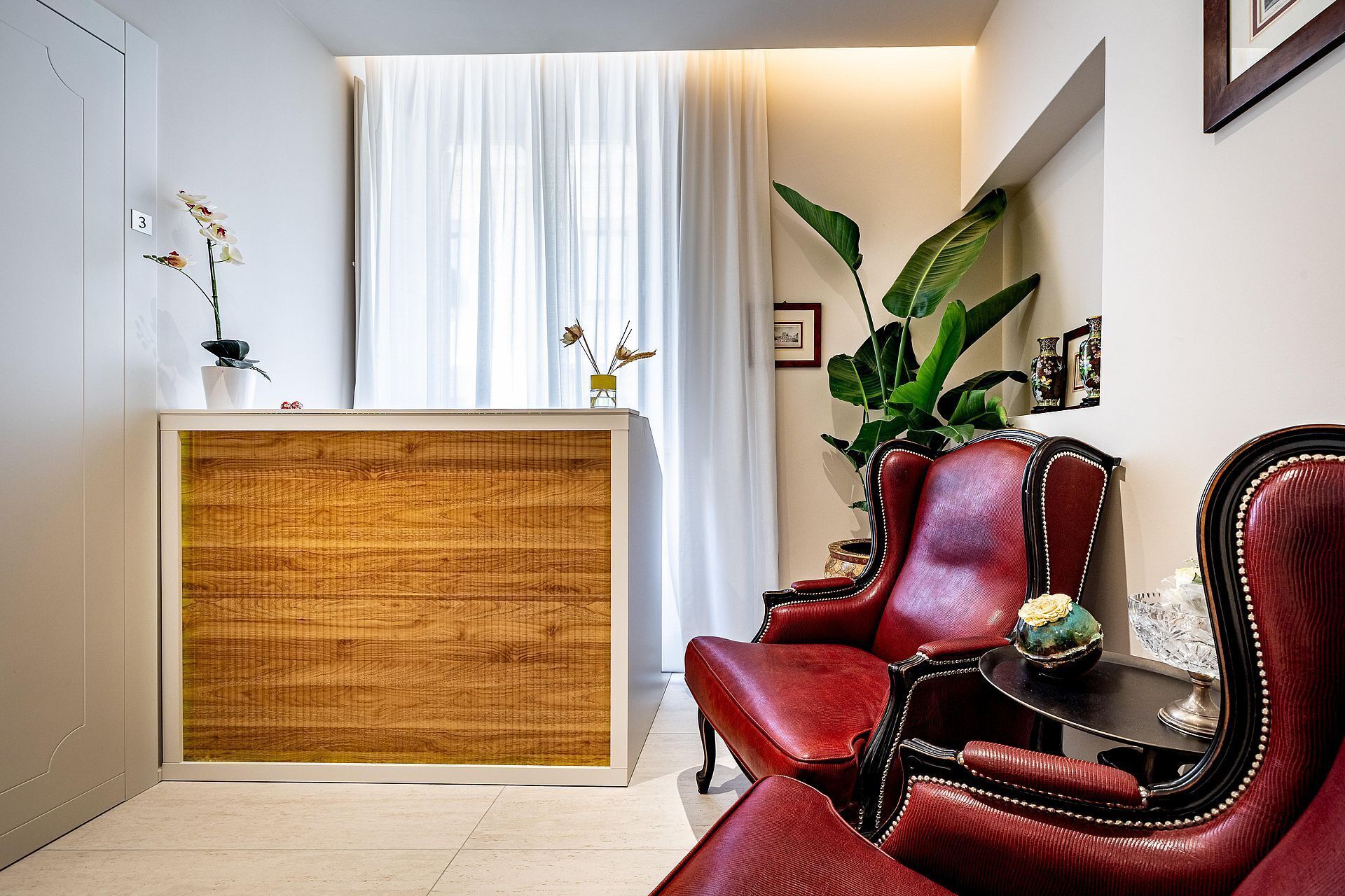 Un comptoir d’accueil avec un panneau en bois, un fauteuil en cuir rouge, une grande plante verte et un tableau encadré au mur.