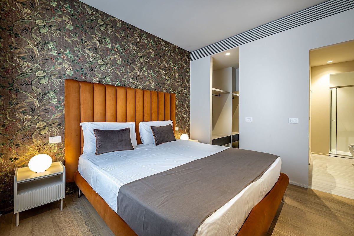Une chambre d’hôtel moderne avec un grand lit, une table de chevet avec une lampe, une salle de bain avec une douche en verre et un mur décoré d’un motif.