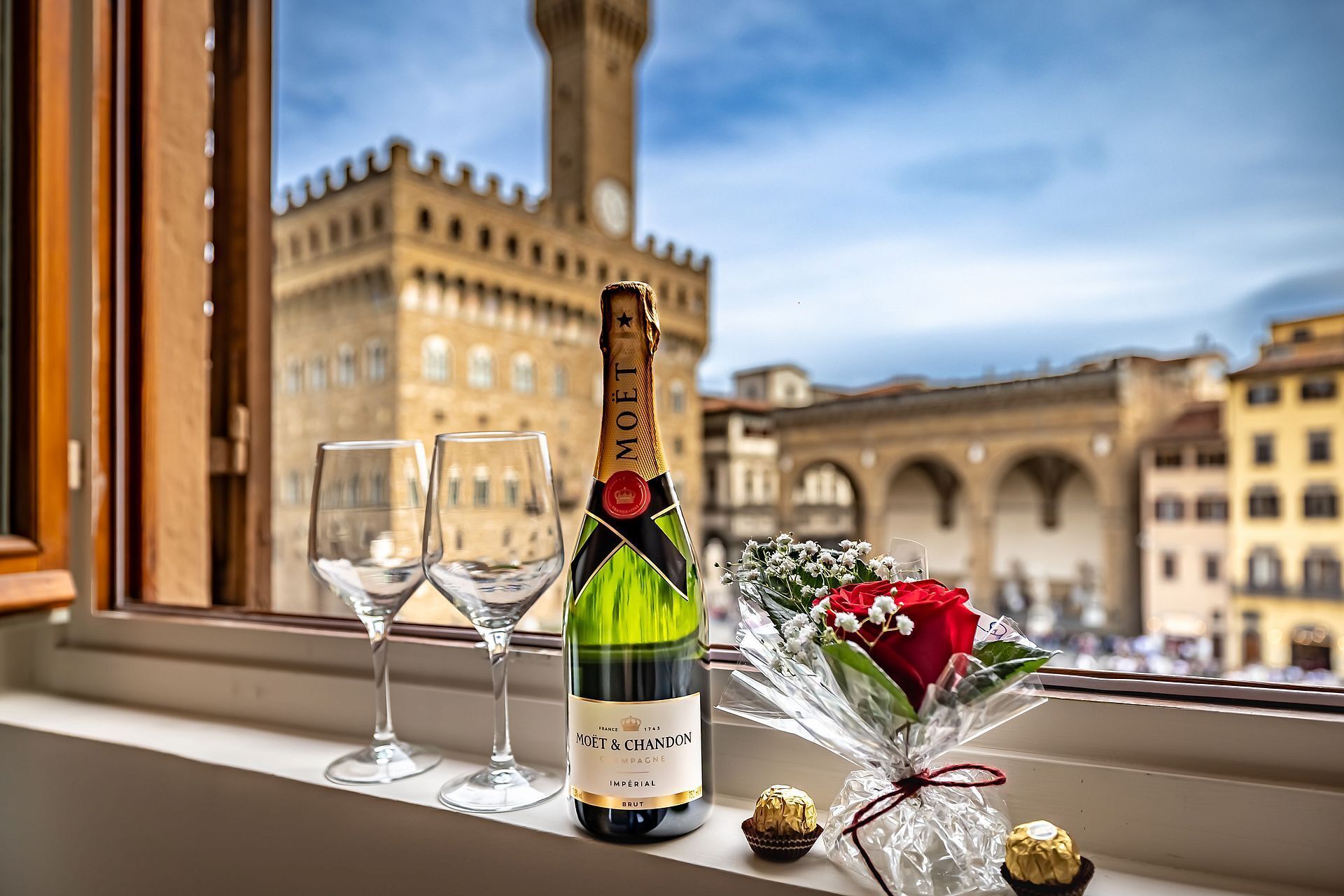 Une bouteille de champagne Moët & Chandon, deux verres vides, un bouquet de fleurs et des chocolats, le tout posé sur un rebord de fenêtre d’où l’on peut admirer des bâtiments d’époque.