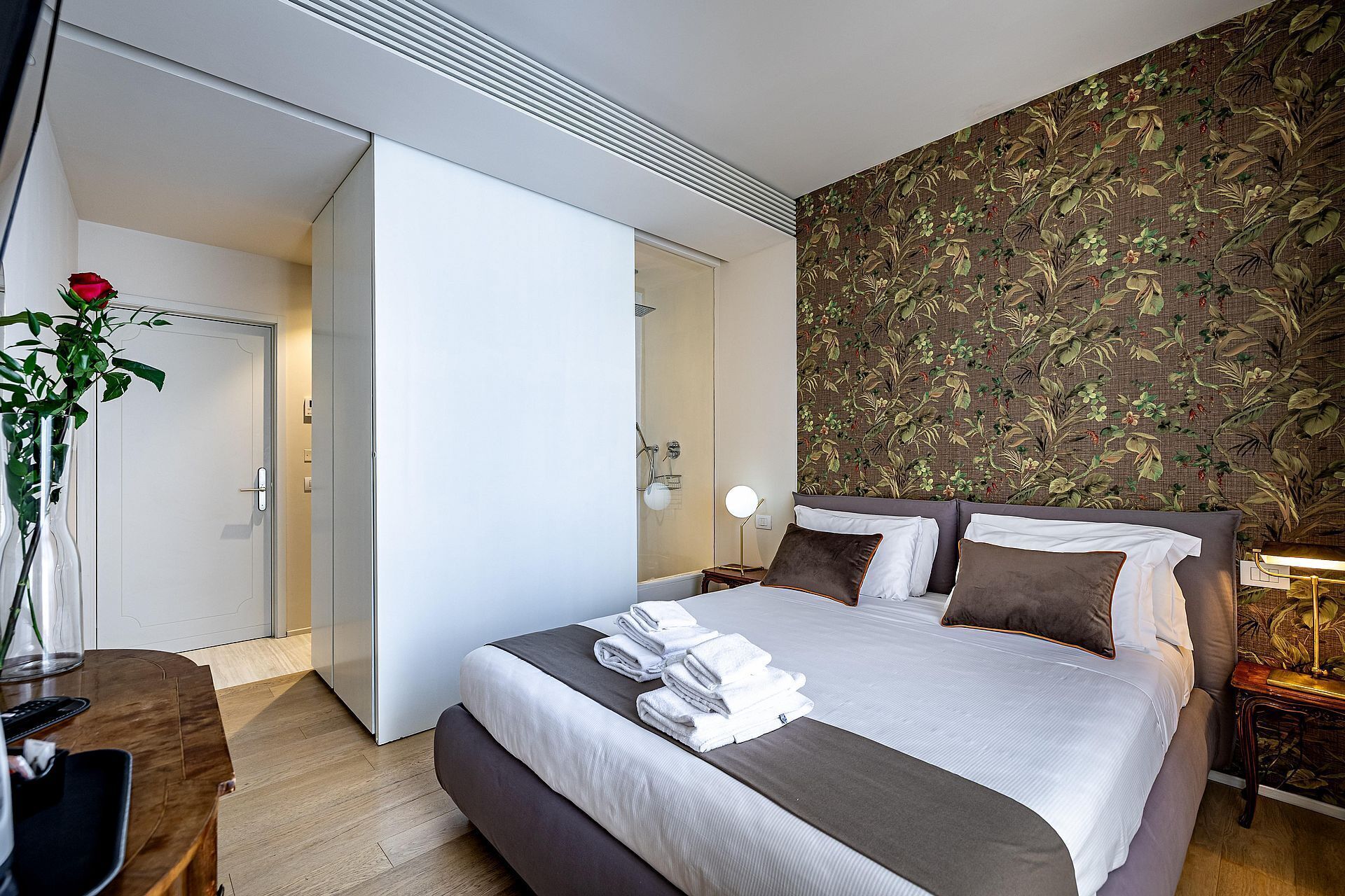 Quarto de hotel com cama, escrivaninha, vaso com planta e área de chuveiro.
