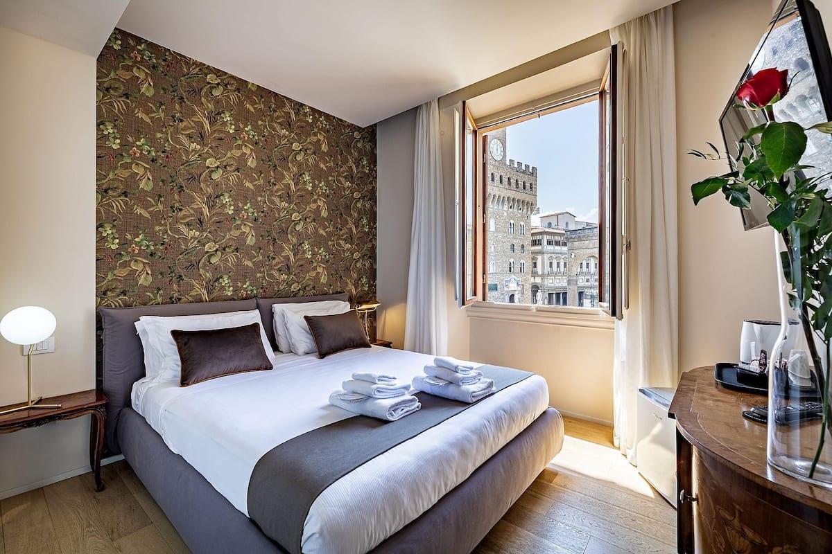 Une chambre d’hôtel avec un lit, une table de chevet, une lampe, une fenêtre donnant sur un bâtiment historique et un vase avec une rose.