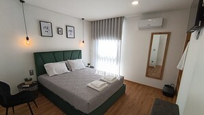 A modern bedroom with a green headboard, a bed with white linens, a small side table with a plant, a chair, a mirror, and an air conditioning unit.