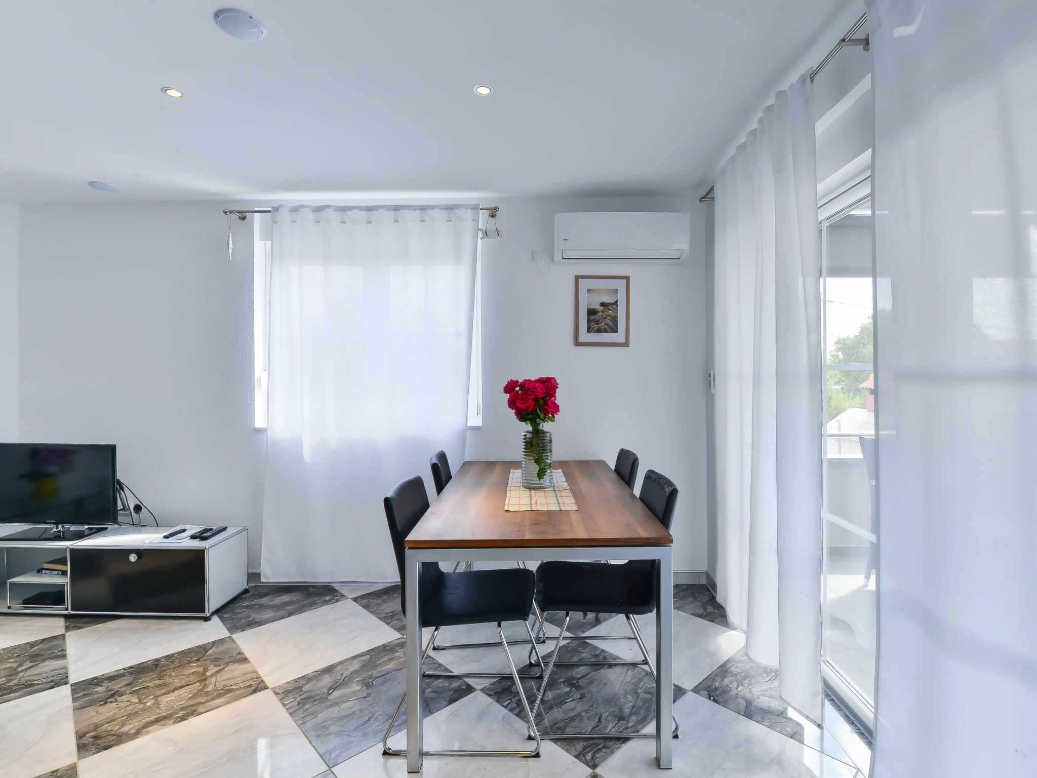 Un espace repas moderne avec une table en bois, des chaises noires, un téléviseur et un vase rempli de fleurs rouges.