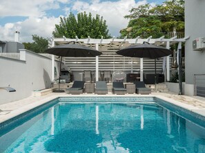 Ein Swimmingpool mit Liegestühlen und Sonnenschirmen, umgeben von einer weißen Pergola und viel Grün.