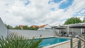 Piscina con plataforma de piedra, rodeada por un muro blanco y una pérgola con sombrillas.