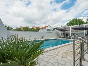 Ein Schwimmbecken mit einer Steinterrasse, umgeben von einer weißen Mauer und einer Pergola mit Sonnenschirmen.