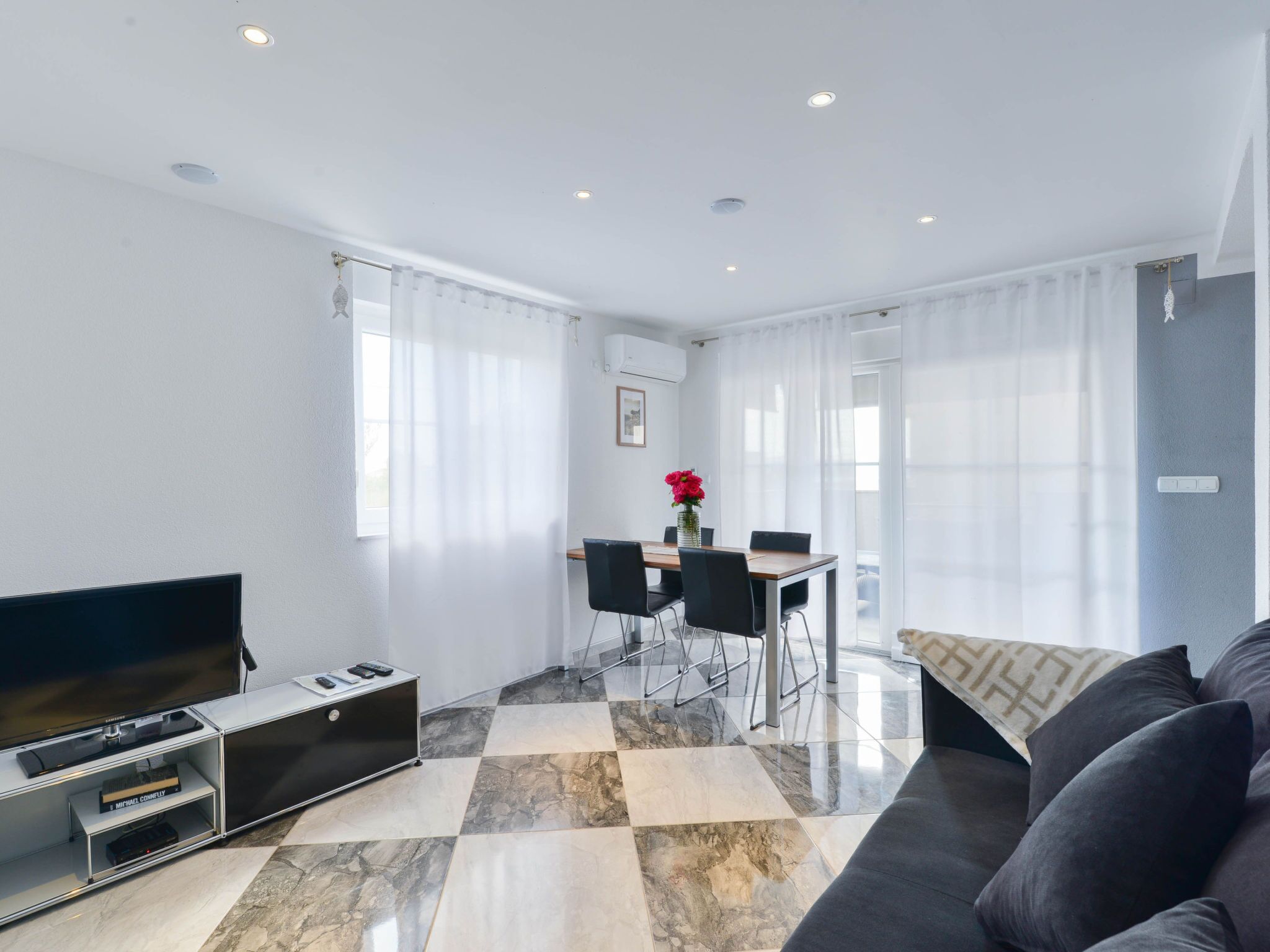 Una sala moderna con un televisor de pantalla plana, un sofá, una mesa de comedor con sillas y un ventanal con cortinas translúcidas.