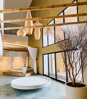 A modern interior with a central seating area, a large potted tree, and a high ceiling with wooden beams and hanging light fixtures. - ID Vida Urbana  (Goiânia)