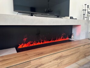 En moderne stue med et fladskærms-TV monteret på en hvid væg, trægulv og en indbygget pejs med synlige flammer.