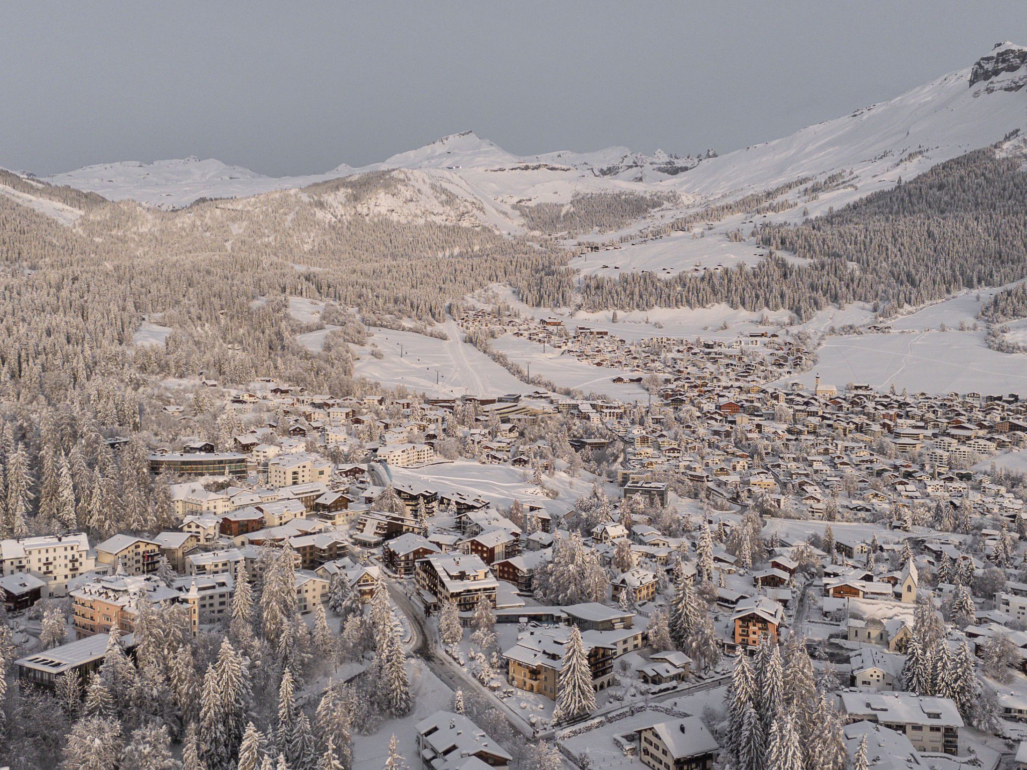 A snow-covered mountain town with buildings clustered around a central area, surrounded by forested slopes and mountains.