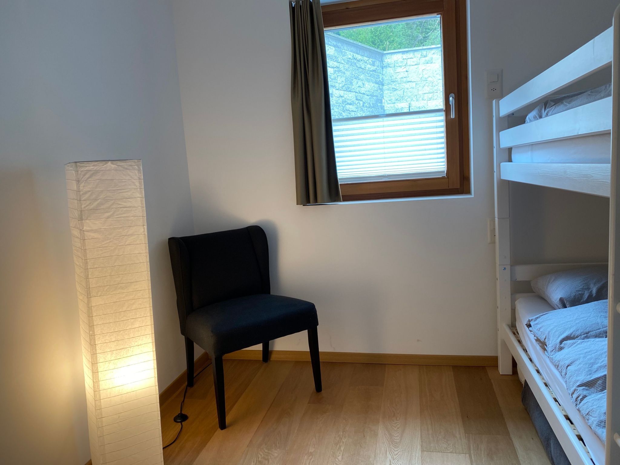 A small room with a bunk bed, a chair, a window with blinds, and a wall-mounted lamp.