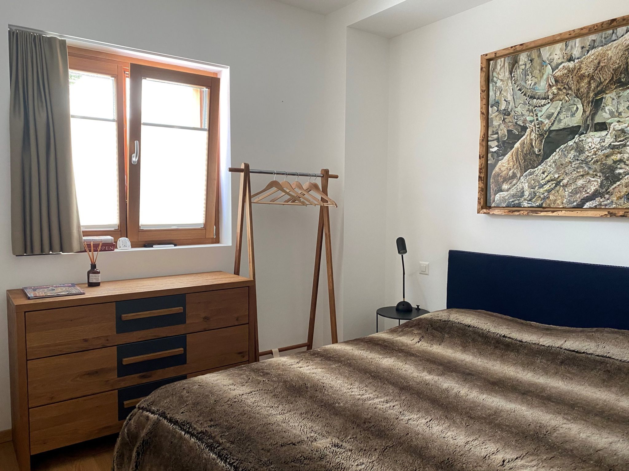 Une chambre avec une commode en bois, un lit recouvert d’une couverture marron, une fenêtre avec des rideaux et un tableau accroché au mur.