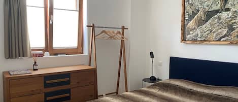 A bedroom with a wooden dresser, a bed with a brown blanket, a window with curtains, and a painting on the wall.