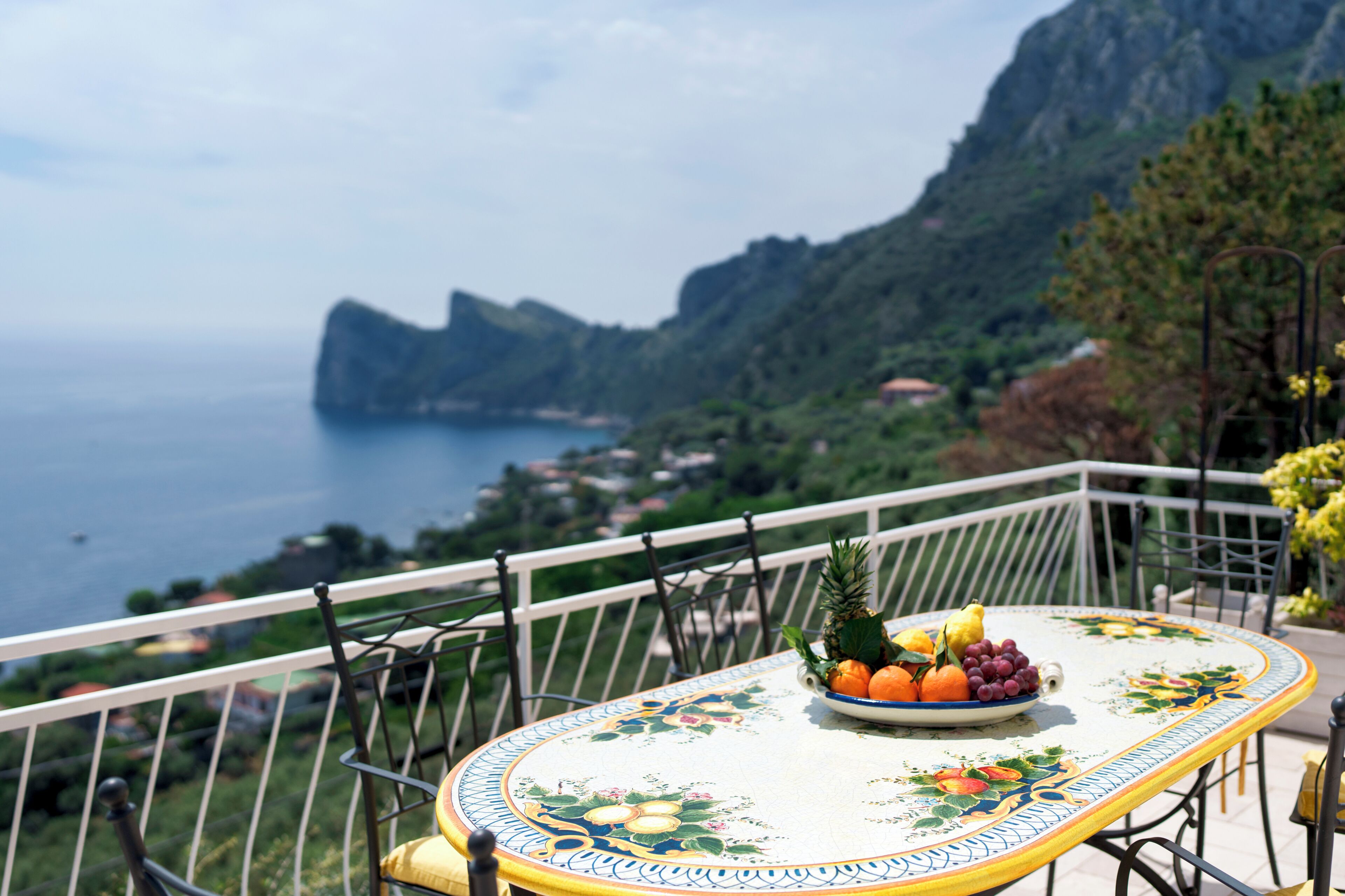 A table with a fruit bowl on a balcony overlooking a coastal landscape.