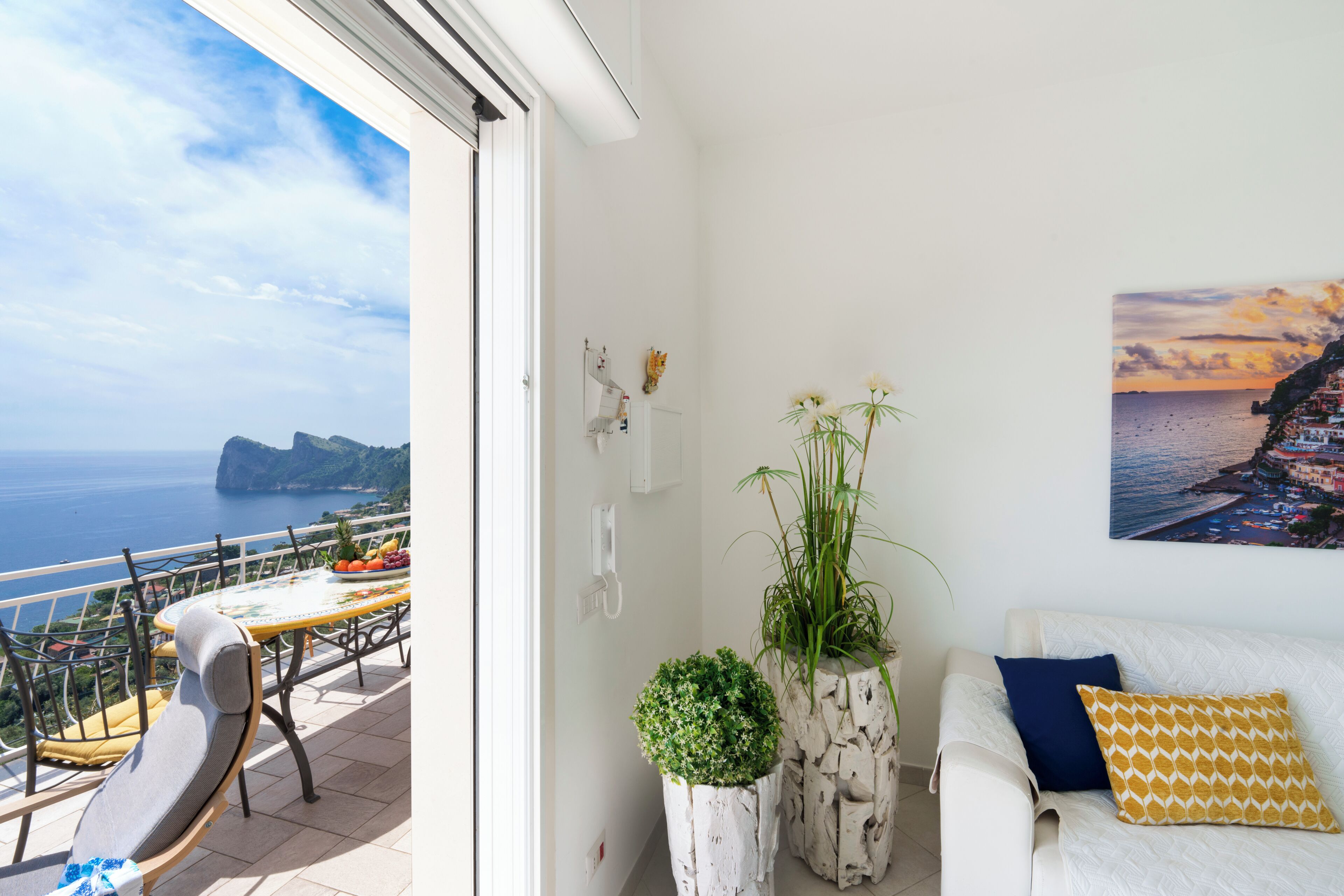 Een balkon met een tafel en stoelen, een witte bank met kussens, een bloempot en een schilderij van een kustlandschap.
