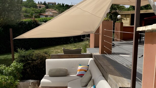 Un espacio al aire libre con luz solar, que incluye un sofá color beige, cojines y una mesa de madera.