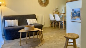 Una sala de estar moderna con un sofá de color oscuro, una mesa de centro de madera, una mesita auxiliar redonda de madera y un taburete pequeño. Hay un espejo montado en la pared, un radiador blanco y una pequeña mesa con sillas.