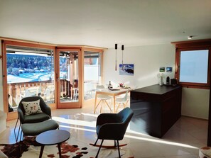 A modern living room with a glass door leading to a balcony, a dining area, and a kitchen with a black countertop. - Fewo Cordial im Casa Erizun, (Flims Dorf), by Interhome (Flims)