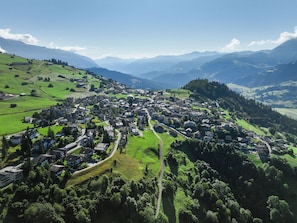 An aerial view of a small town with a central road, surrounded by green hills and mountains.