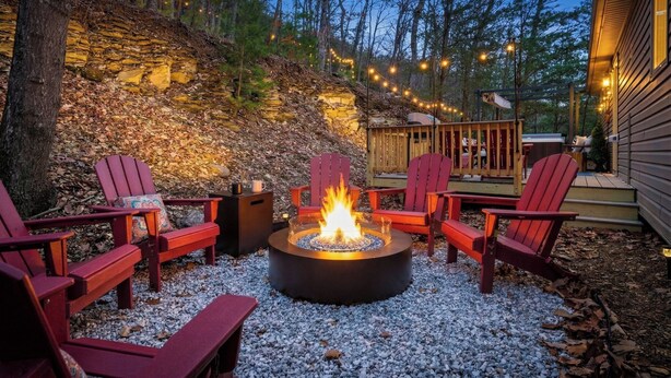 Uma fogueira rodeada por cadeiras Adirondack, um muro de pedra e um deck com luzes de corda.