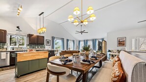 Cozinha moderna com ilha central, mesa de jantar e cadeiras. Há um ventilador de teto, iluminação pendente e vista para o exterior.