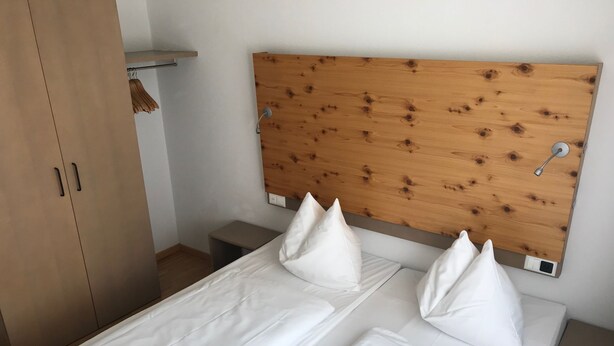 Een bed met witte lakens en twee kussens, een houten hoofdbord, een houten garderobe en een klein nachtkastje.