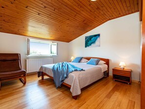 A bedroom with a wooden ceiling, a bed with a blue blanket, a chair, a nightstand with a lamp, and a window with a view. - Apartment house Simoni with pool by Interhome (Privlaka)