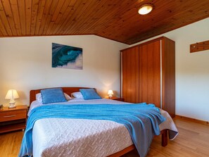 Un dormitorio con techo de madera, cama con ropa de cama azul, mesita de noche con lámpara y armario de madera.
