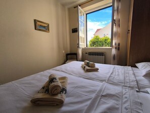 A hotel room with two beds, a window with a view of greenery, a radiator, and a framed picture on the wall. - House Deša with amazing Old Town and sea view (Dubrovnik)