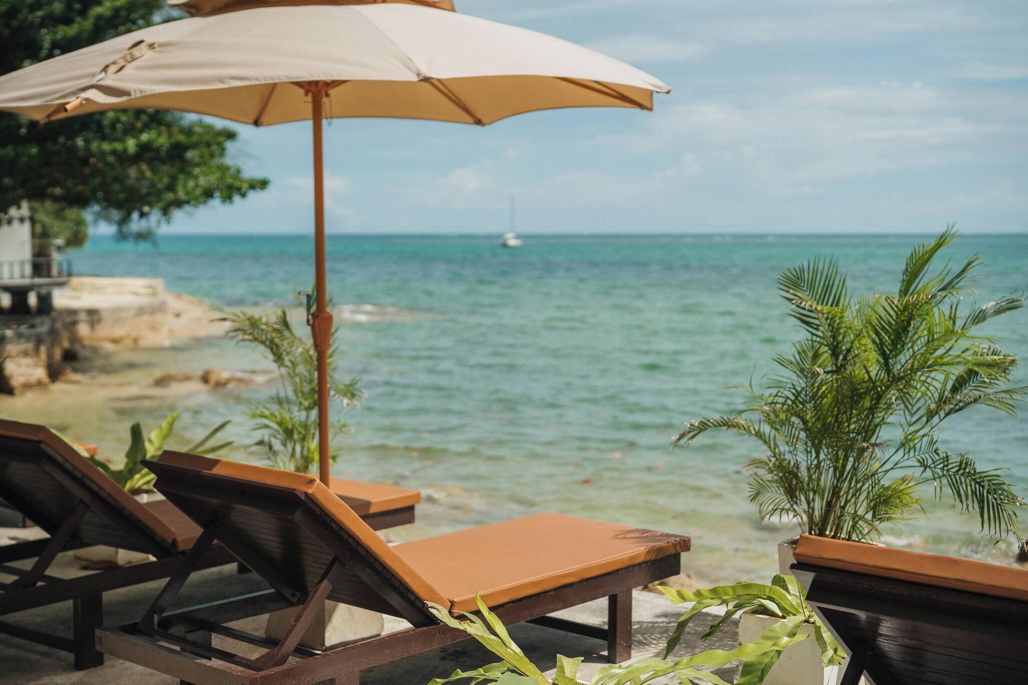A beachside lounge with wooden chairs and a beige umbrella, overlooking a calm sea and a distant sailboat.