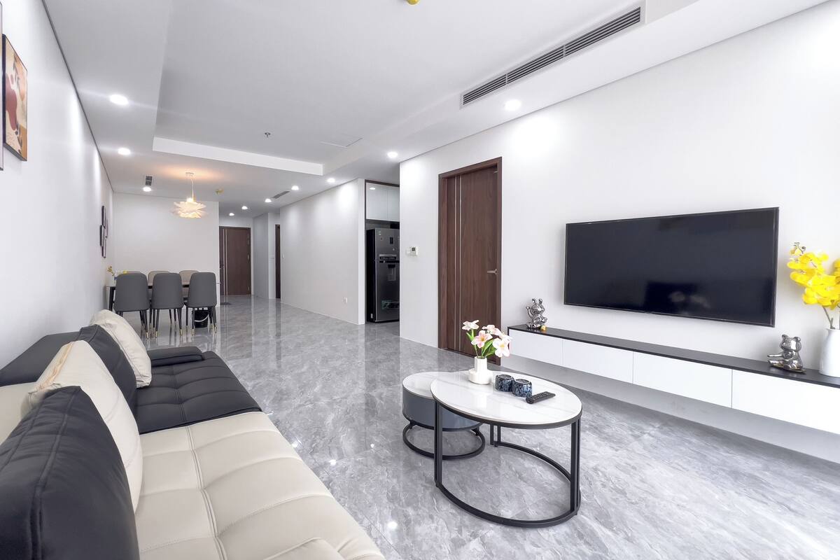 A modern living room with a sectional sofa, a dining area, a flat-screen TV, and a small round table with decorative items.