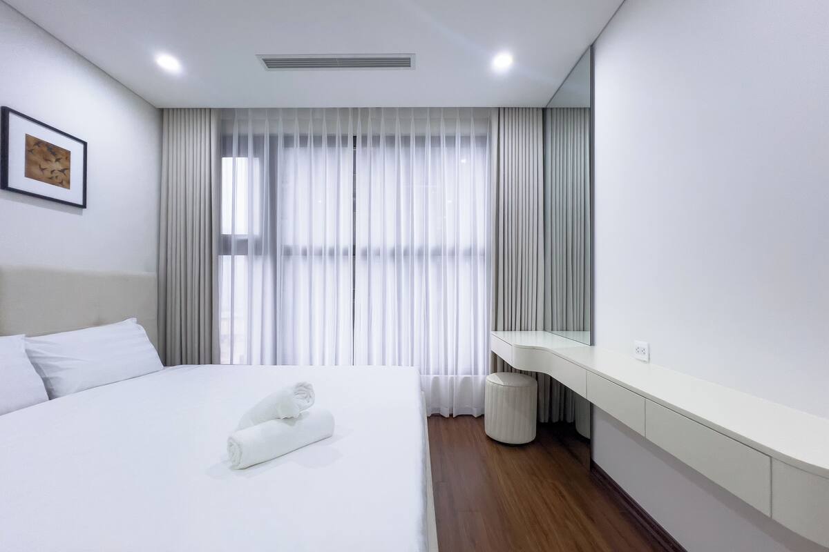 A modern hotel room with a neatly made bed, a large window with sheer curtains, a bedside table, and a mirror.