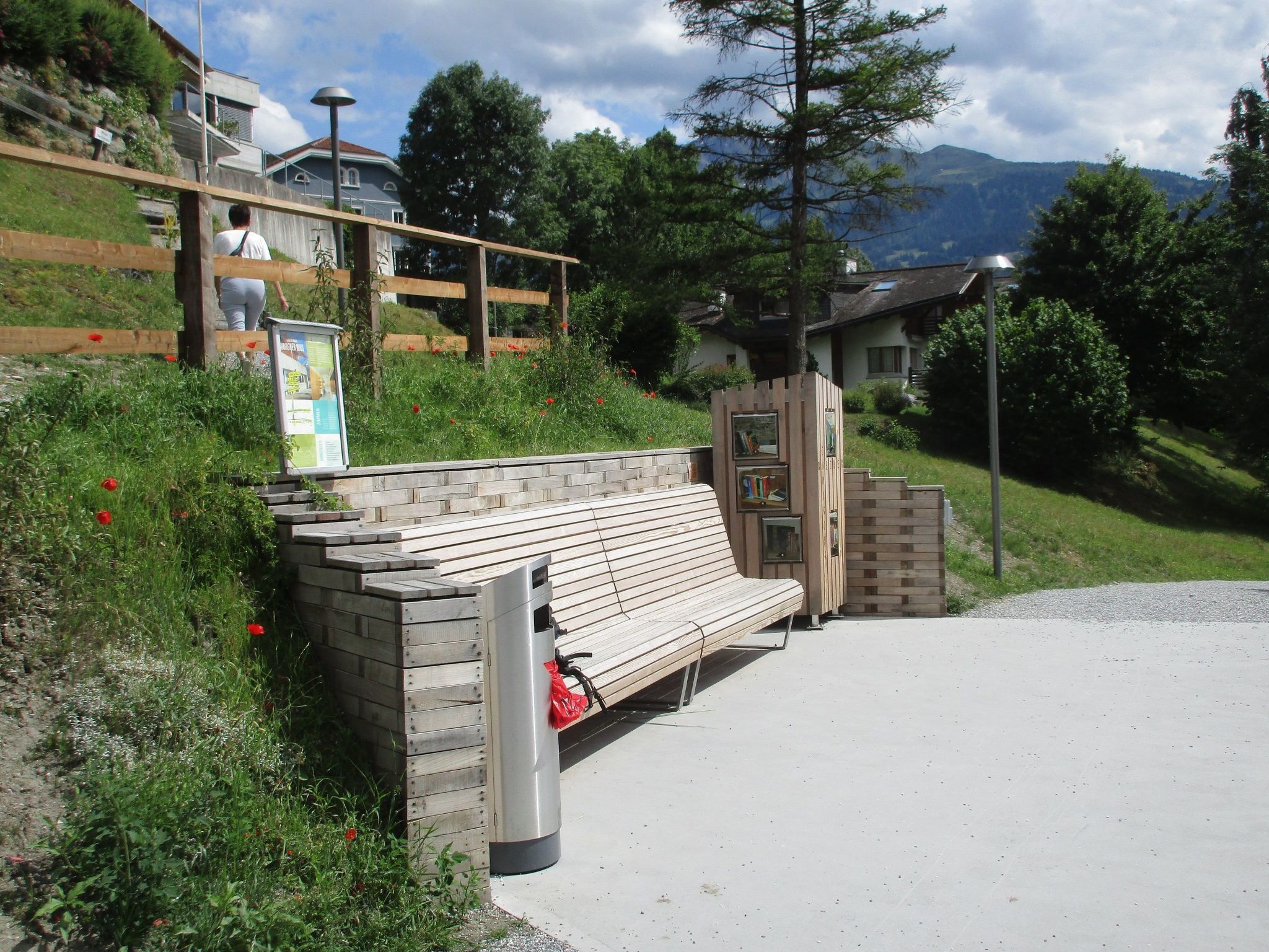 A wooden bench with a metal trash can beside it, set against a backdrop of a grassy area with a mountain in the distance.