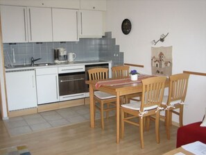 A compact kitchen with white cabinets, a built-in oven, and a coffee maker. There is a dining table with wooden chairs and a red cushion. - La Prada, (Laax Dorf), 20047 by Interhome (Laax)