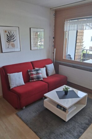 Un canapé rouge agrémenté de coussins blancs et à carreaux, une table basse blanche avec une plante, et des œuvres d’art encadrées accrochées au mur.