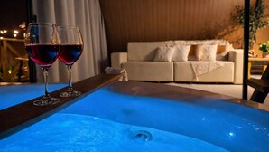 A hot tub with two glasses of red wine, a sofa, and a wooden table in a room with a curtain and a plant. - Cabana com Piscina Aquecida e Spa (Palhoça)