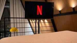 A hotel room with a bed, a TV mounted on the wall displaying the Netflix logo, a window with curtains, and a yellow lamp. - Cabana com Piscina Aquecida e Spa (Palhoça)