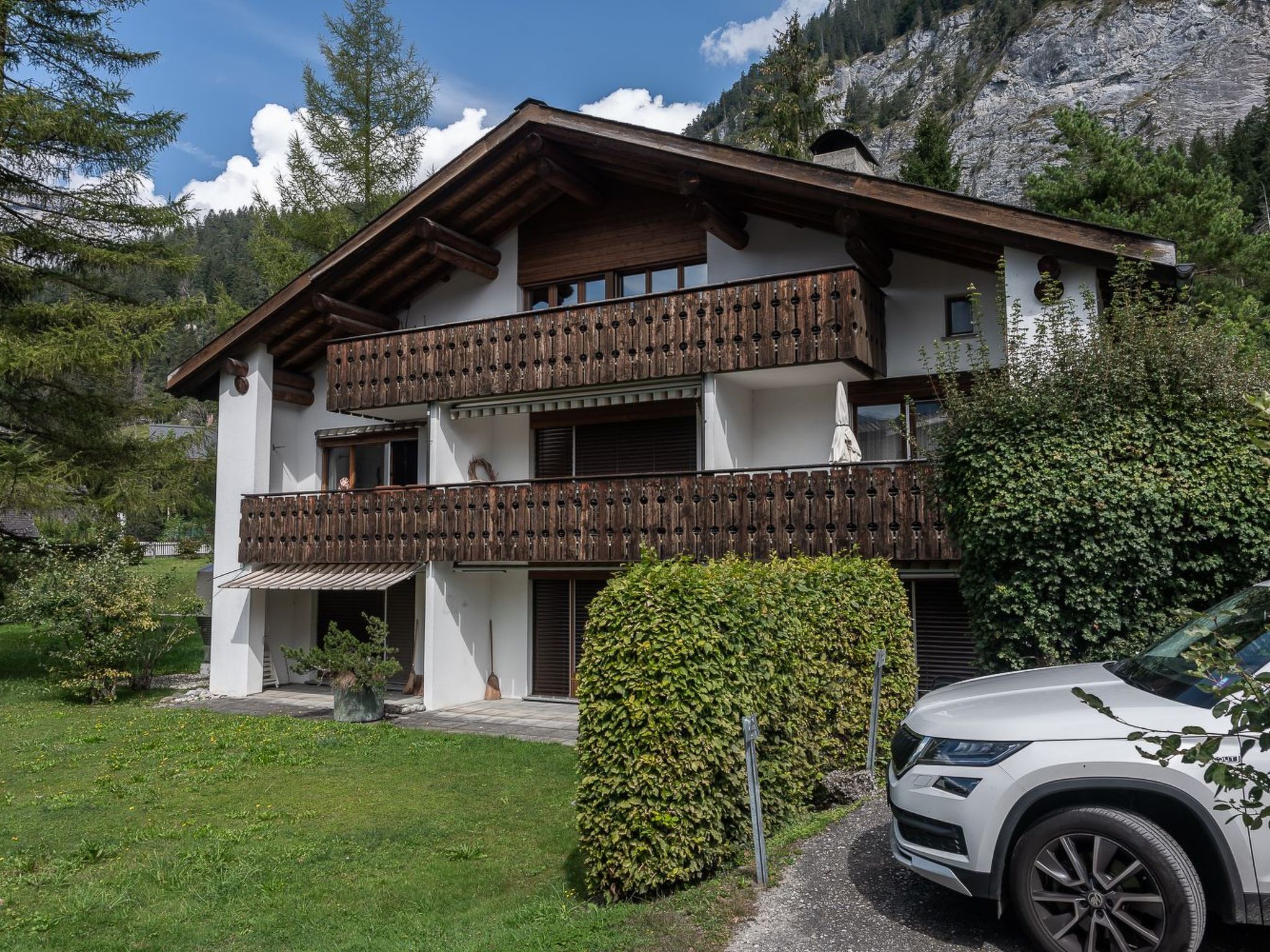 A two-story chalet with wooden balconies, a car parked in front, and a mountainous backdrop.