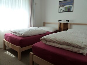 A room with two single beds, each with a wooden frame and a red mattress. There is a small wooden shelf on the wall above the beds. - Surrieven / Zwicker, (Laax Dorf), 257 by Interhome (Laax)