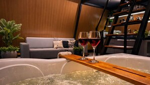A hot tub with two glasses of red wine on a wooden tray, a grey sofa with a cushion, and a wooden shelf in the background. - Cabana Industrial com Hidro e Piscina Aquecida (Palhoça)